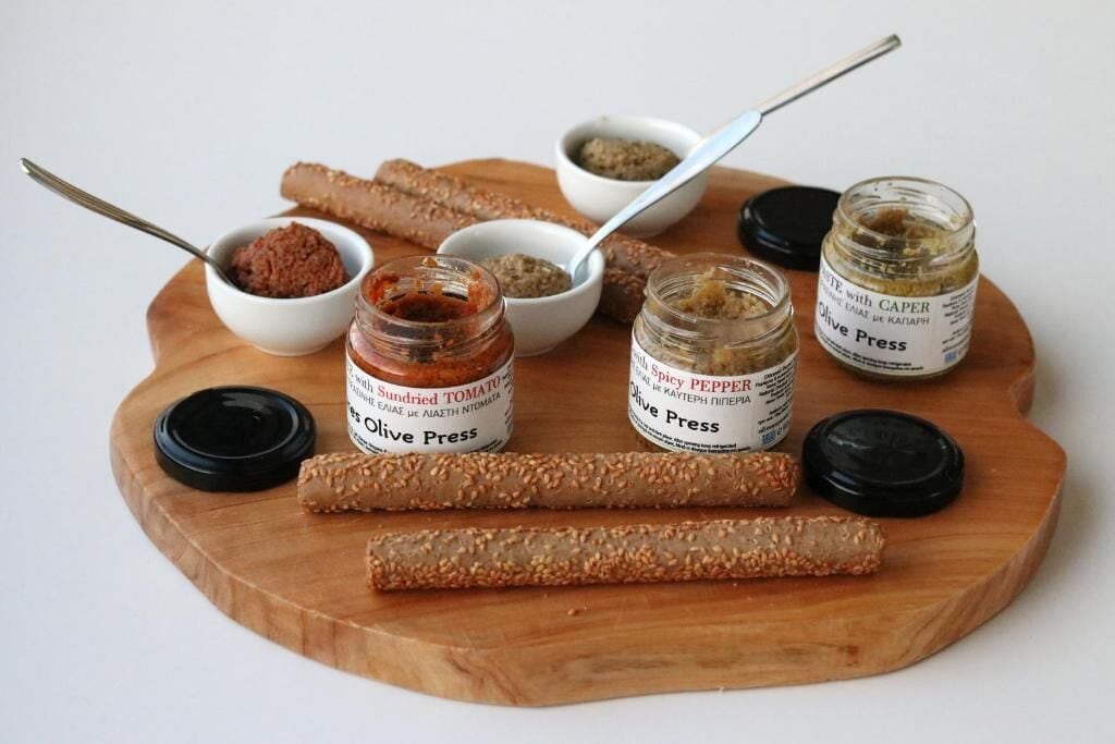 A wooden board with jars of olive press condiments and two wooden sticks.