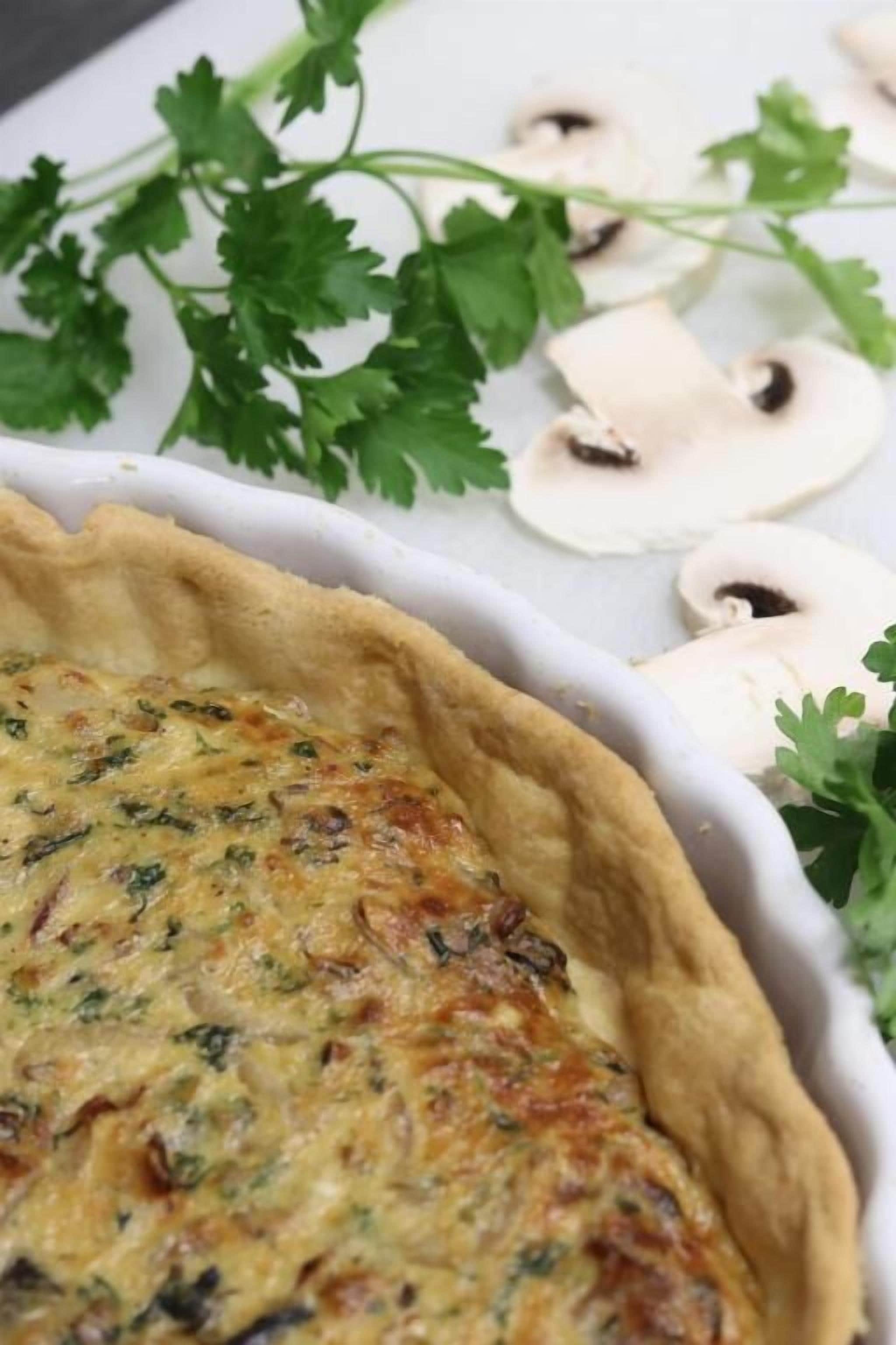 A quiche in a white ceramic dish with a fluted edge, surrounded by parsley and sliced mushrooms.