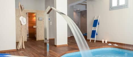 Una piscina interior moderna con una fuente de agua cristalina, suelo de madera y una toalla azul en una silla.