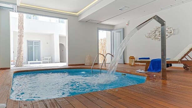 An indoor swimming pool with a wooden deck, a water fountain, and a lounge area with a blue towel. - Grotta Hotel (Naxos)