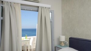 A hotel room with a bed, a nightstand with a lamp, and a view of the ocean through a window with curtains. - Grotta Hotel (Naxos)