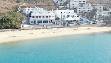 Un edificio costero con una playa al frente, una carretera con automóviles estacionados y un paisaje montañoso en el fondo.