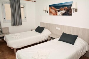 Habitación con dos camas individuales, cada una con una almohada y una toalla doblada. Se aprecia una ventana con cortina gris, un cuadro colgado en la pared de una playa y una lámpara de pared.