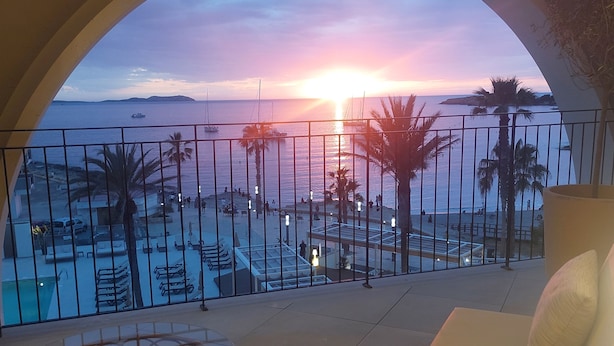 A balcony with a view of the sunset over the sea, palm trees, and a pool area. - Blau Park (Sant Antoni de Portmany)