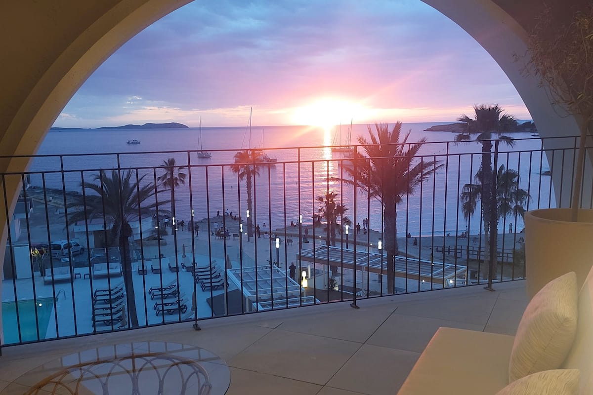 Ein Balkon mit Blick auf den Sonnenuntergang über dem Meer, Palmen und einen Poolbereich.