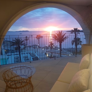 Un balcón con vistas a la puesta de sol sobre el mar, palmeras y una zona de piscina.