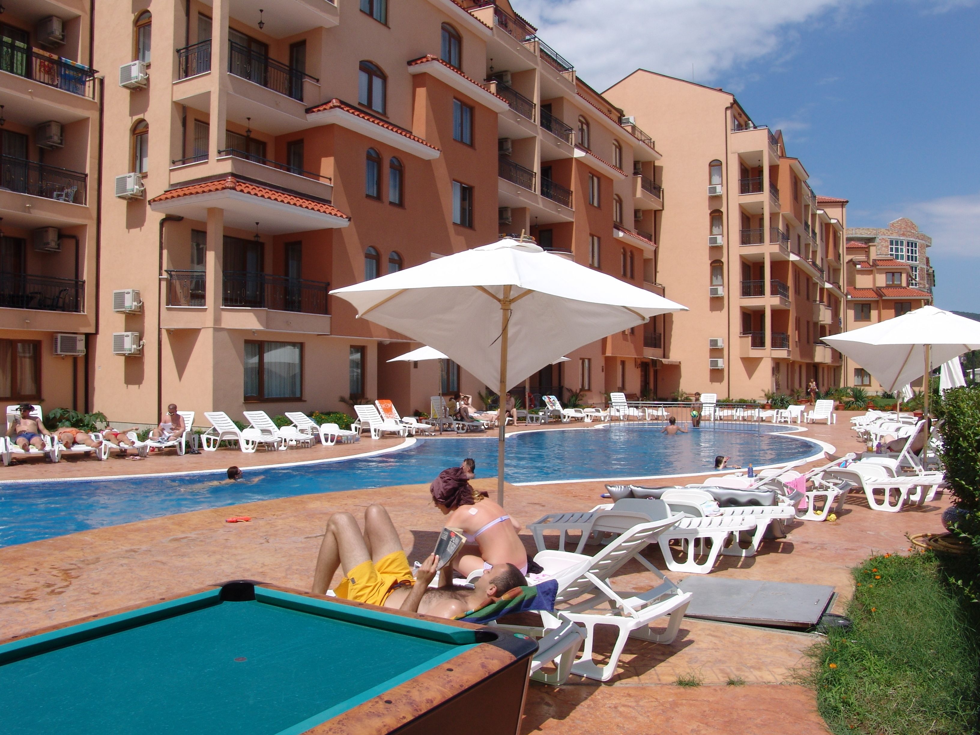 A poolside area with lounge chairs and umbrellas, surrounded by multi-story buildings with balconies.