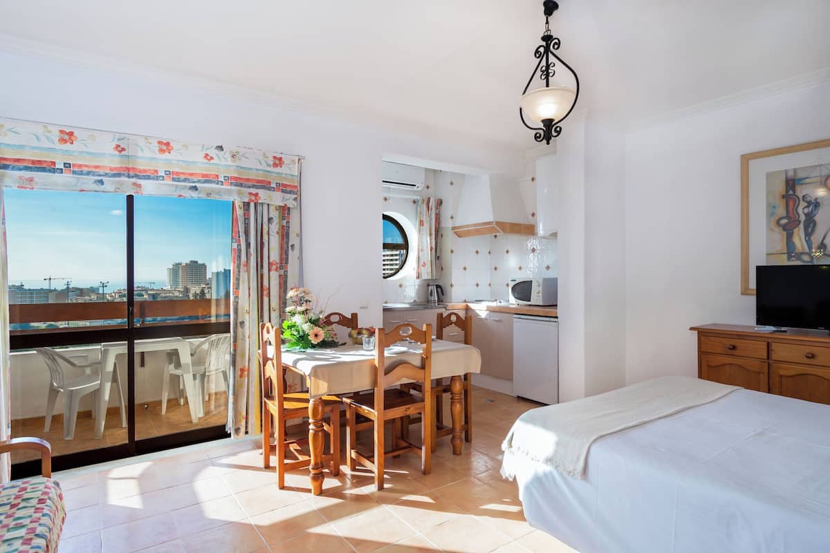 Ein Schlafzimmer mit Bett, Fernseher, einer Essecke mit Stühlen und Tisch sowie einem Balkon mit Blick auf die Stadt.