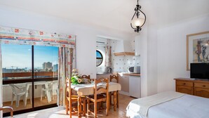Et soveværelse med seng, fjernsyn, spiseplads med stole og bord, samt en balkon med udsigt over byen.