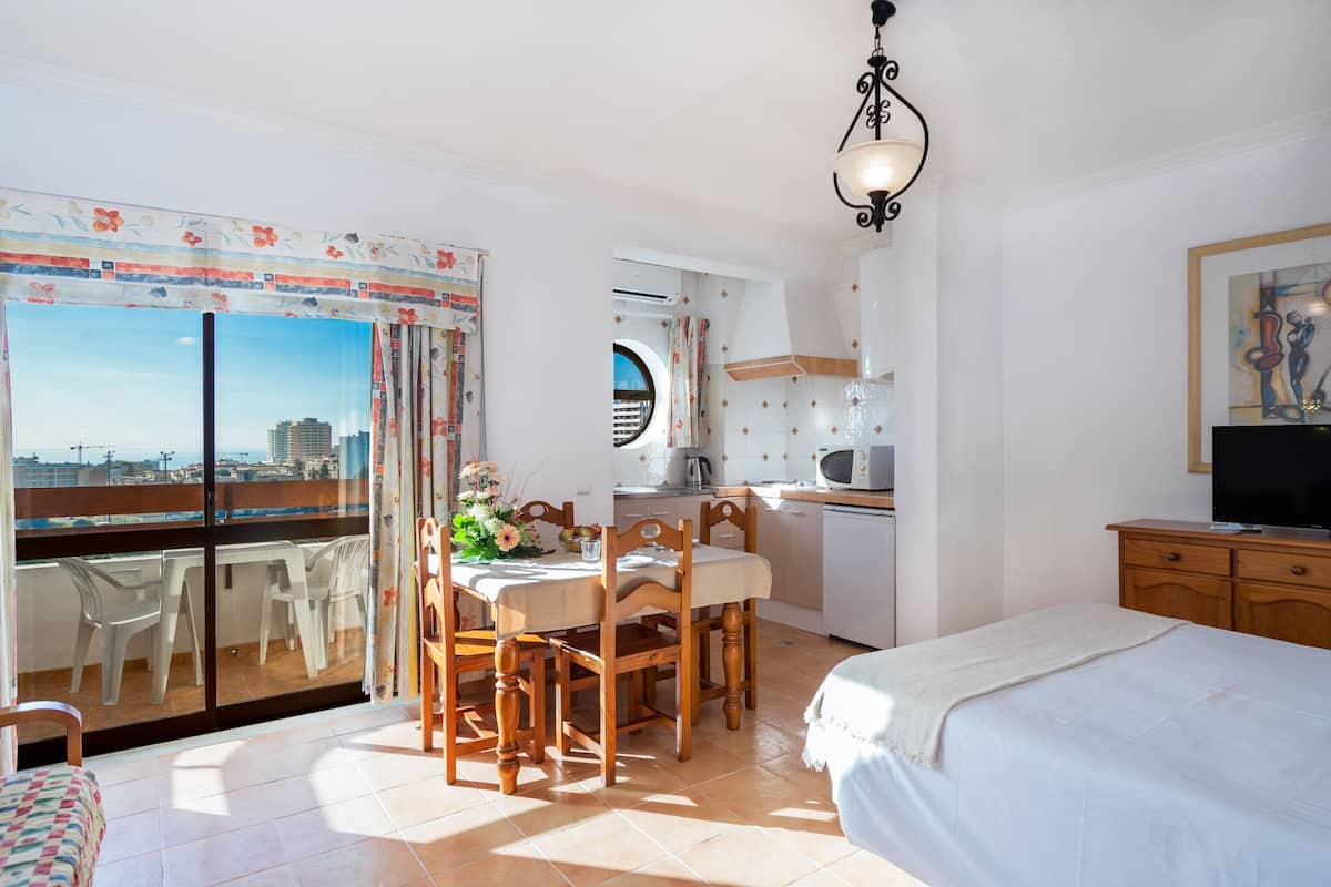 Ein Schlafzimmer mit Bett, Fernseher, einer Essecke mit Stühlen und Tisch sowie einem Balkon mit Blick auf die Stadt.