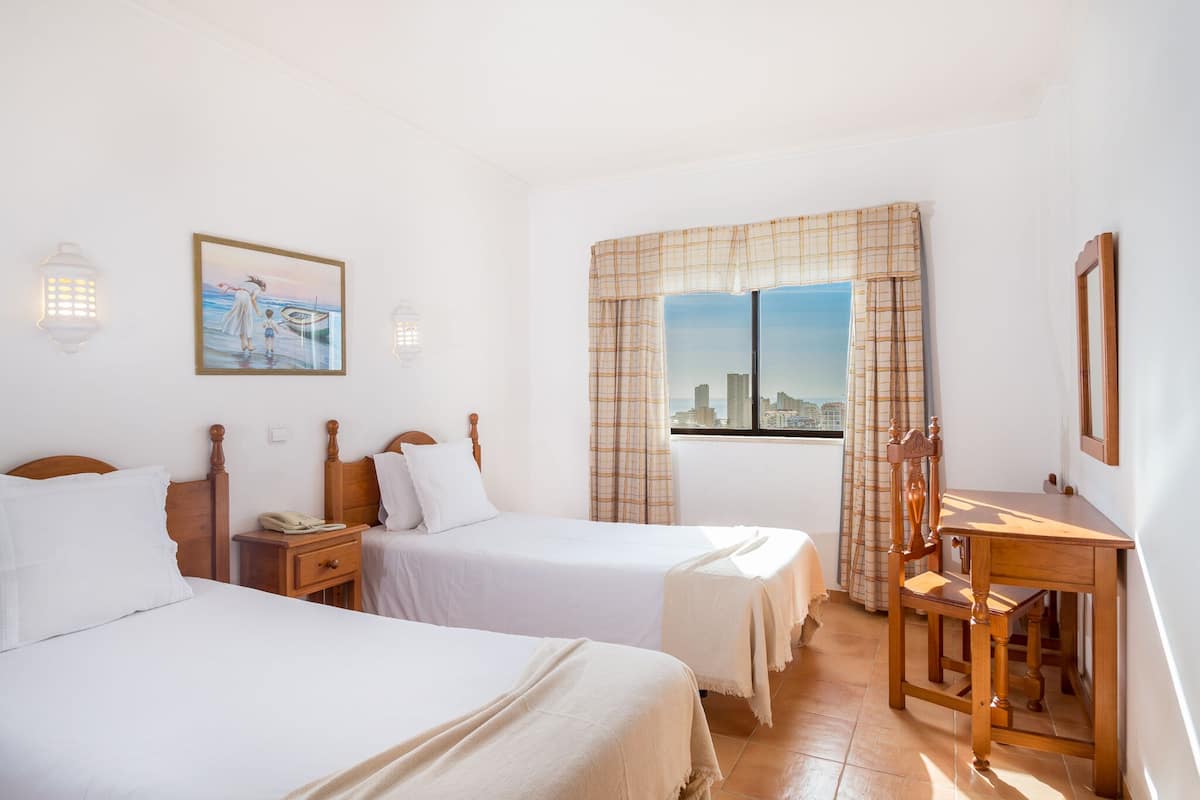 Ein Hotelzimmer mit zwei Betten, einem Schreibtisch, einem Stuhl, einem Fenster mit Vorhängen, Blick auf die Stadt und Wandleuchten.
