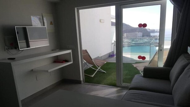A modern room with a sofa, a TV on a stand, a small table, and a sliding glass door leading to a balcony with a pool view. - Puerto Principe Apartments (PUERTO RICO)