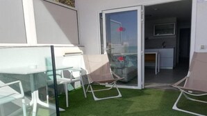 A modern outdoor patio with a glass table and chairs, a cushioned chair, and a sliding glass door leading to a kitchen area. - Puerto Principe Apartments (PUERTO RICO)