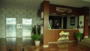 Un comptoir d’accueil avec une enseigne indiquant « Réception » et « Hôtel de plage ». Le hall d’entrée comprend deux ascenseurs et des plantes en pot.
