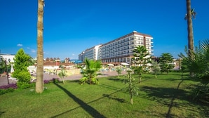 A hotel with a large building, a grassy area with palm trees, and a clear blue sky. - Eftalia Splash Resort (Alanya)