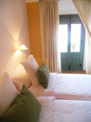 Cama doble con almohadas blancas y verdes, una lámpara de pared y vistas a una puerta y cortinas azules.