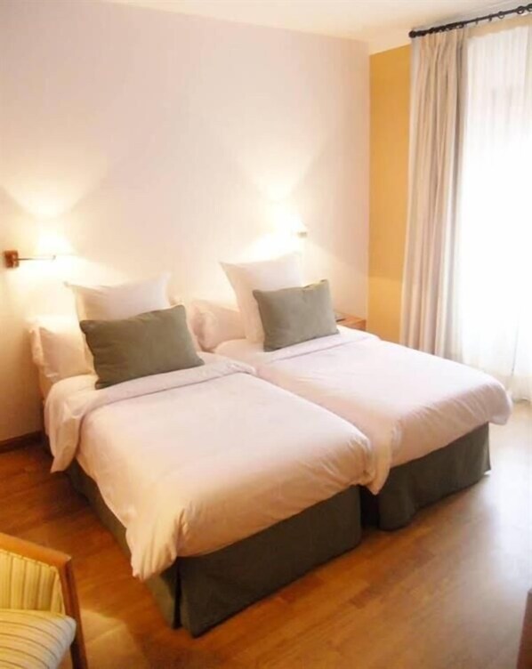 A double bed with two pillows, a nightstand, and a chair in a room with wooden flooring and a curtain on the window. - AHC Palacio Coria (Coria)
