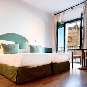 Une chambre d’hôtel avec un grand lit, une vue sur un bâtiment en pierre par une fenêtre ouverte et un balcon avec une chaise.