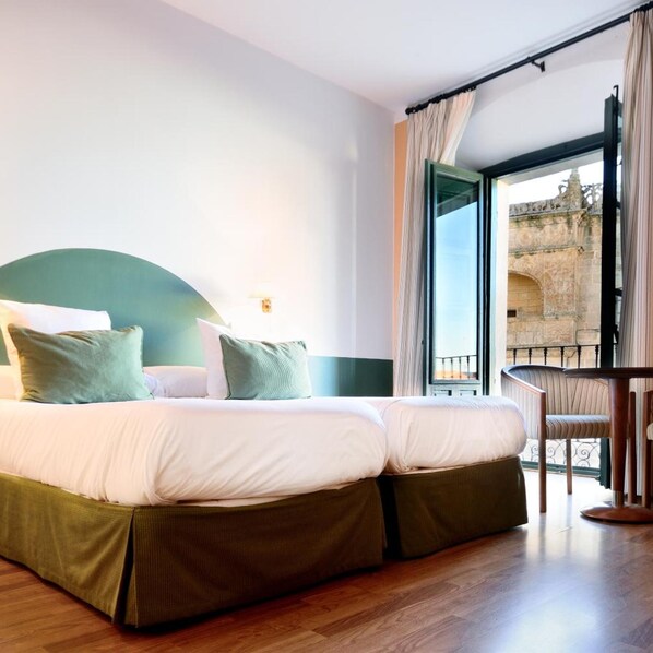 A hotel room with a large bed, a view of a stone building through an open window, and a balcony with a chair. - AHC Palacio Coria (Coria)