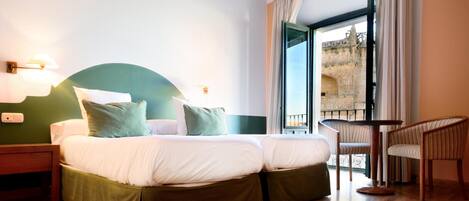 A hotel room with a double bed, two bedside tables, a chair, and a balcony with a view of a stone building.