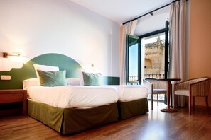 Une chambre d’hôtel avec un lit double, deux tables de chevet, une chaise et un balcon donnant sur un bâtiment en pierre.