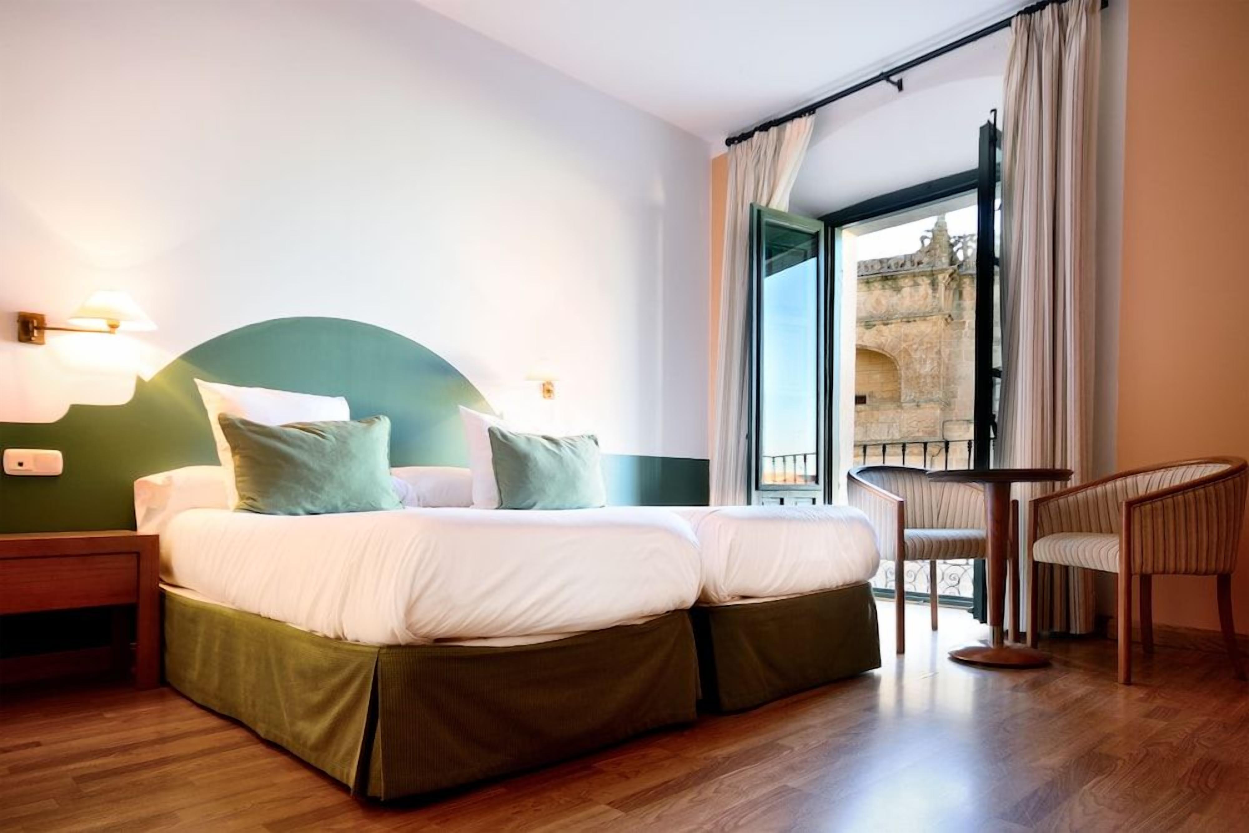 A hotel room with a bed, a chair, and a balcony with a view of a historic building.