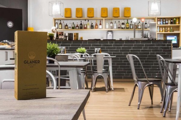 L'intérieur d'un café moderne présente une table en bois, des chaises en métal et un mur en briques. On y trouve des étagères remplies de bouteilles et un présentoir à menus.