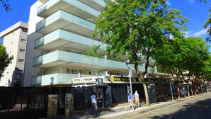 Un edificio moderno de varios pisos, con una tienda de tatuajes y perforaciones en la planta baja, y un área cercada con árboles.