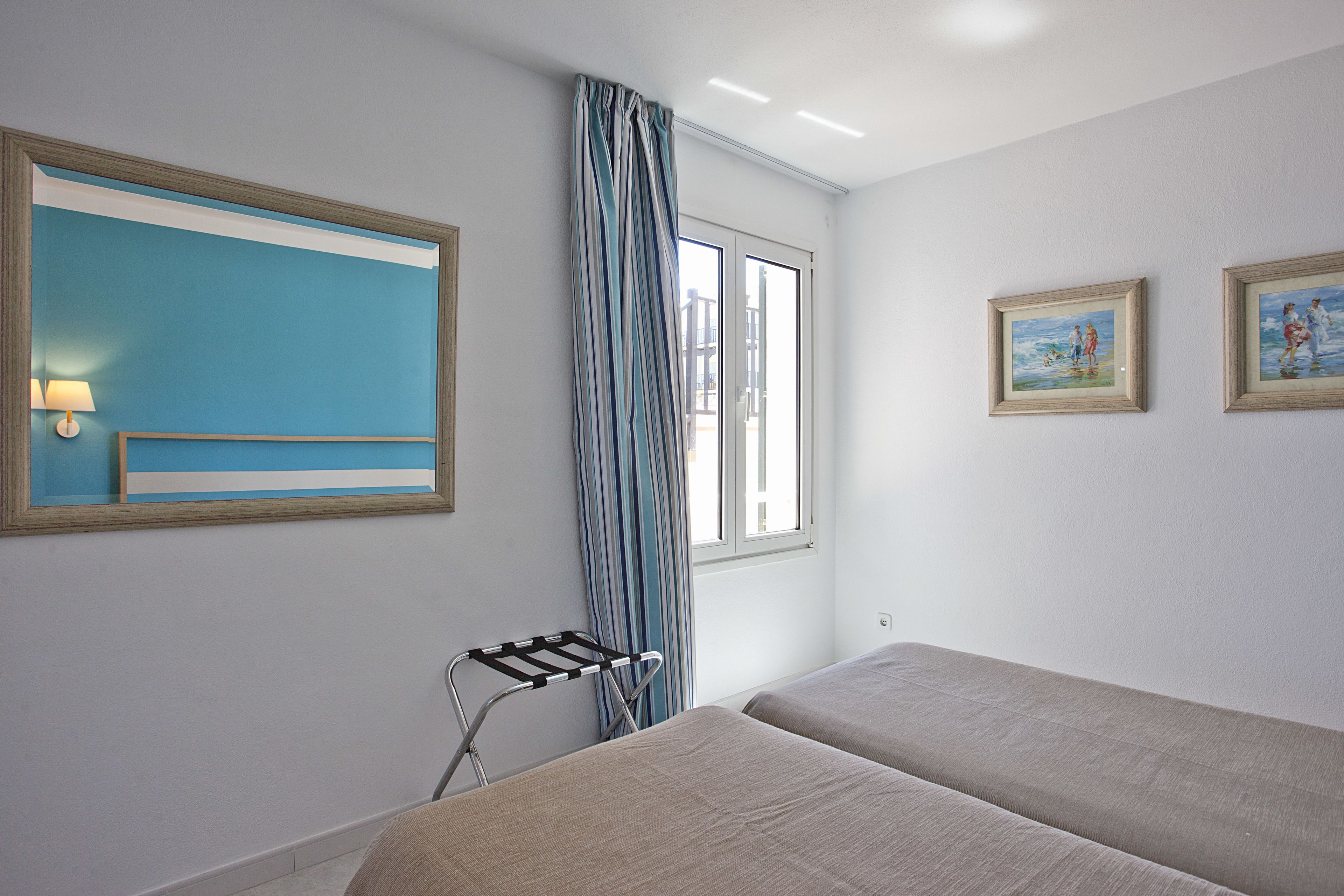 Habitación de hotel con dos camas, una ventana con cortinas, una lámpara de pared y dos cuadros enmarcados.