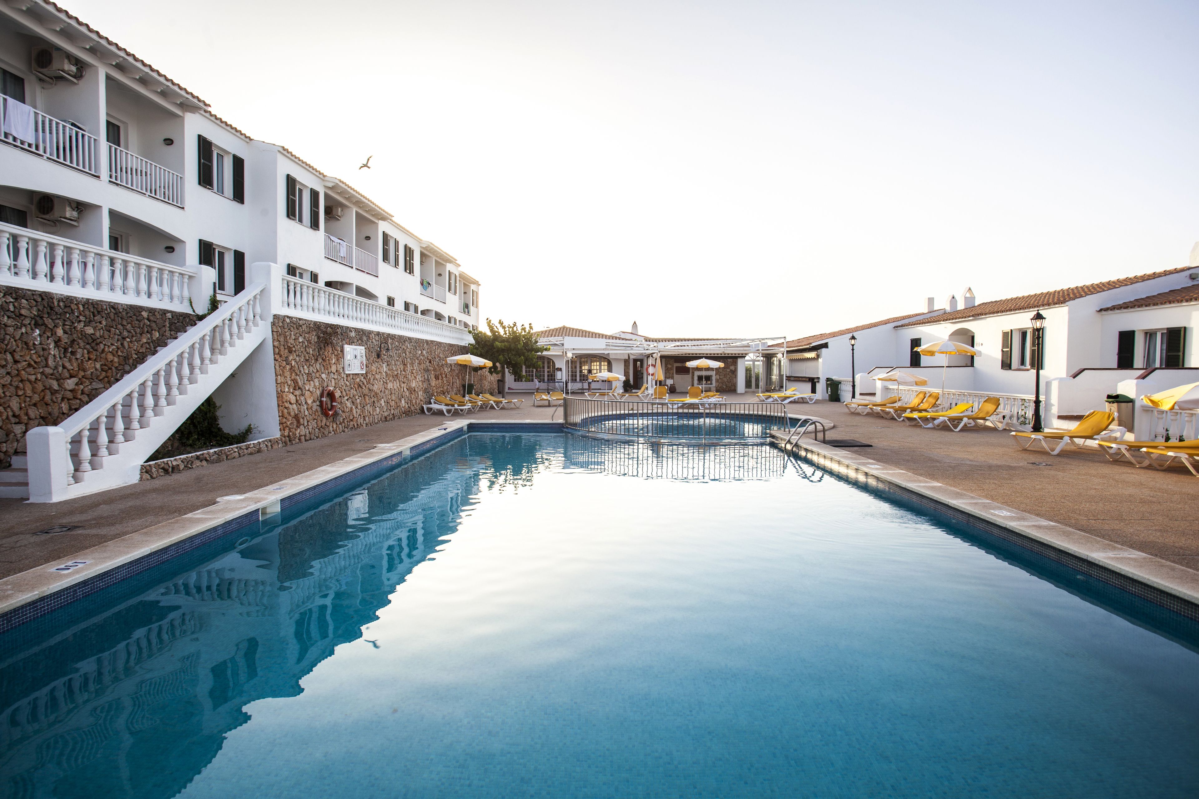 A swimming pool with sun loungers and a staircase leading to a building with balconies.