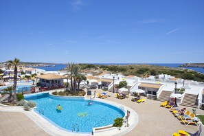 A pool area with a clear view of the sea, lounge chairs, and a building with a tiled roof. - Aparthotel Seth Isla Paraiso (Mercadal)