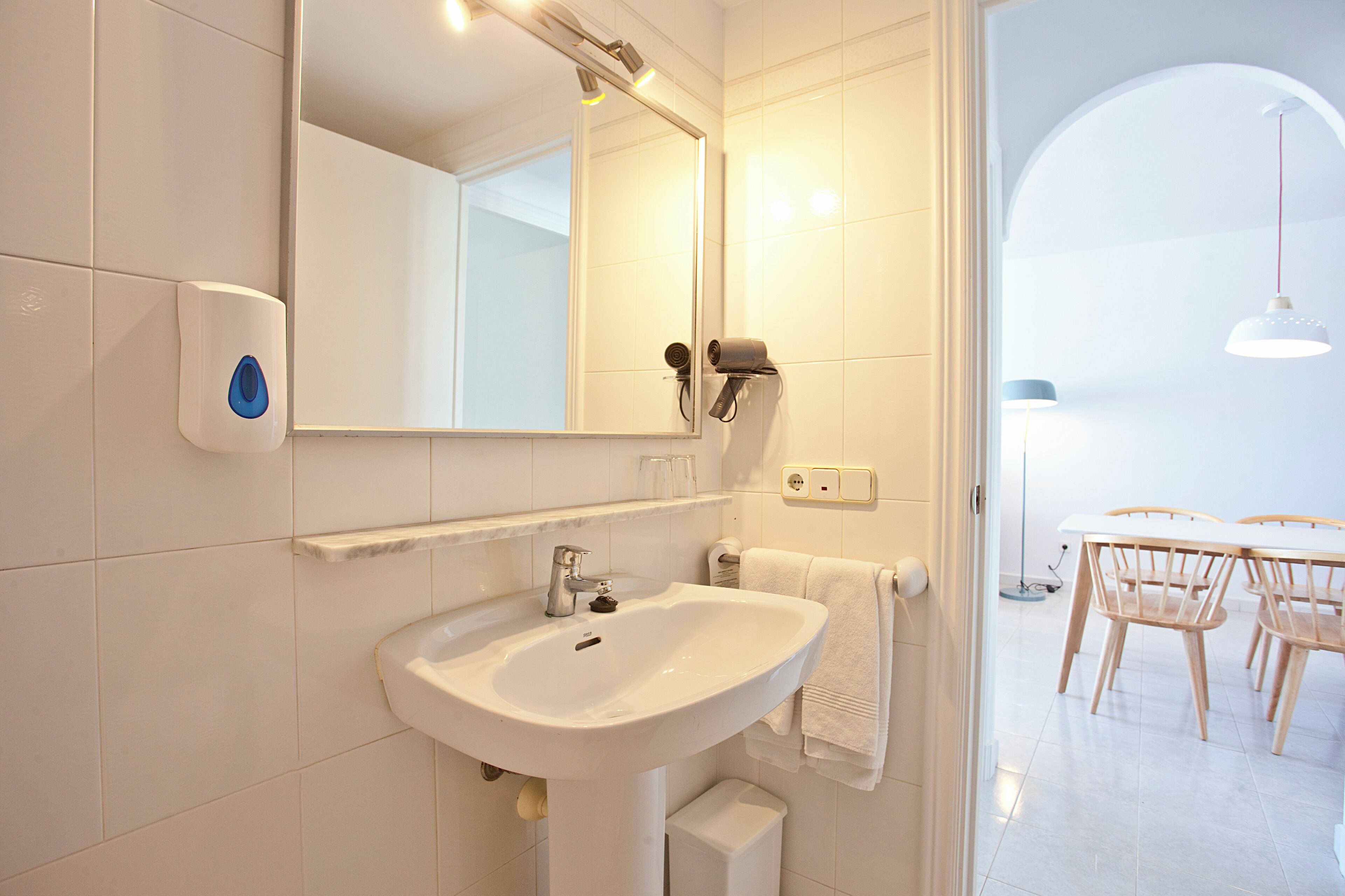 Un baño con lavamanos blanco, espejo y barra para toallas. Hay un secador de pelo y un dispensador de jabón en la pared.