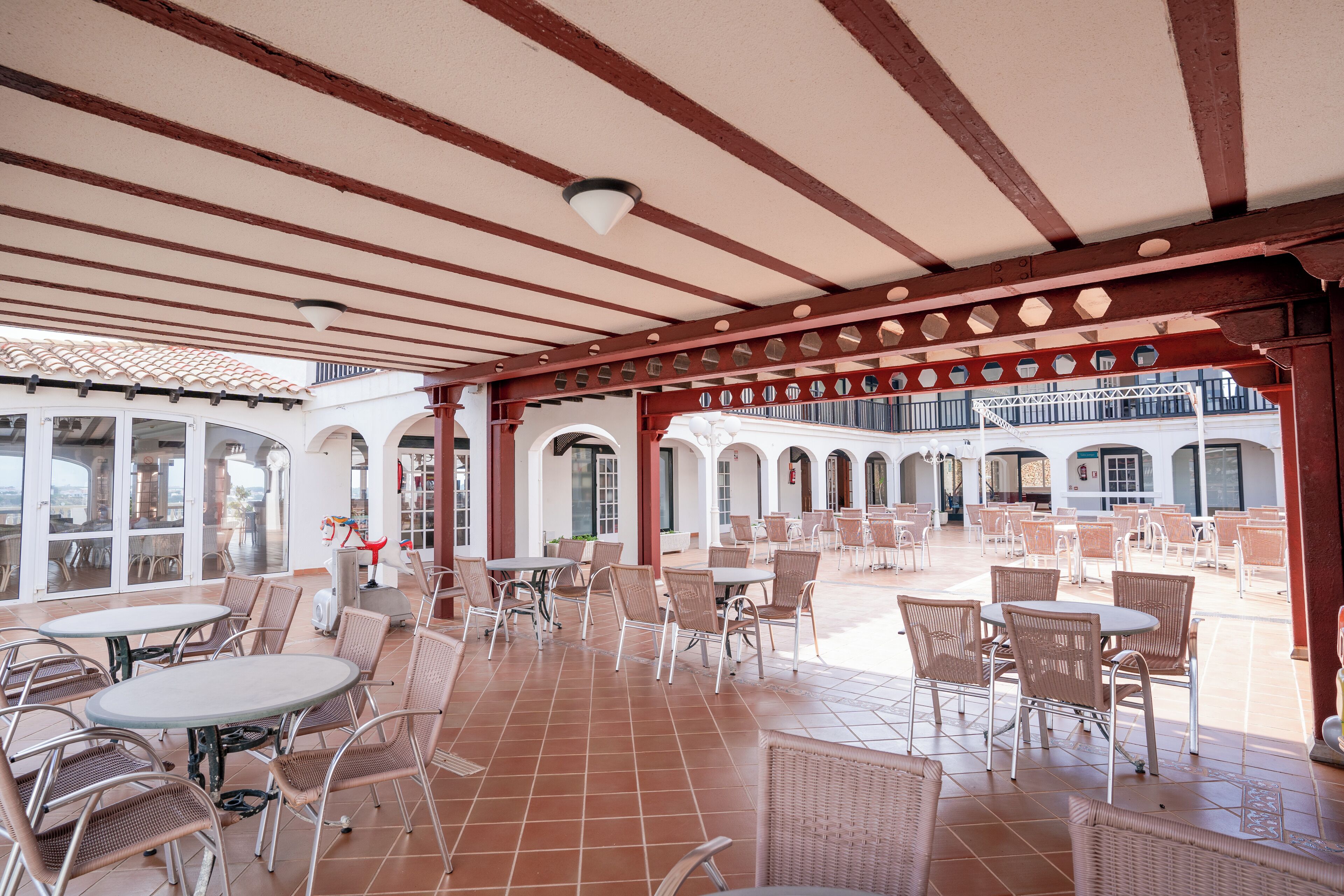 An outdoor patio area with tables and chairs, a tiled floor, and a covered section with a red beam structure.