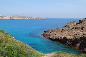 A coastal landscape with clear blue waters, rocky terrain, and sparse vegetation.