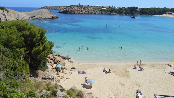 A beach with clear turquoise water, rocky cliffs, and a few people swimming and sunbathing. - Aparthotel Seth Isla Paraiso (Mercadal)