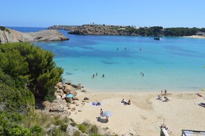 A beach with clear turquoise water, rocky cliffs, and a few people swimming and sunbathing. - Aparthotel Seth Isla Paraiso (Mercadal)