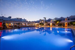 Una piscina con superficie azul cristalina, rodeada de edificios hoteleros con balcones y palmeras.