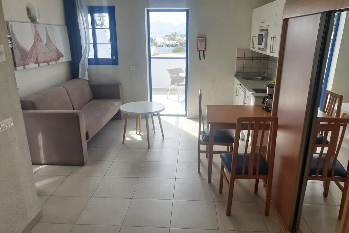 Un espacio habitable compacto con sofá, una mesita redonda y una mesa de comedor con sillas. Se observa una zona de cocina con microondas y fregadero.