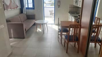 A compact living space with a sofa, a small round table, and a dining table with chairs. A kitchen area with a microwave and sink is visible.