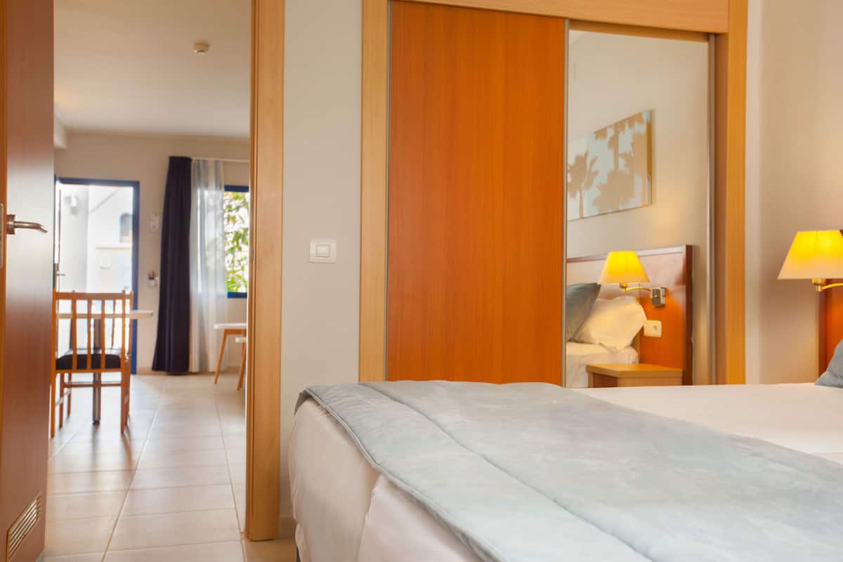 Una habitación de hotel con cama, lámparas de noche, un armario de madera, una silla y un escritorio.