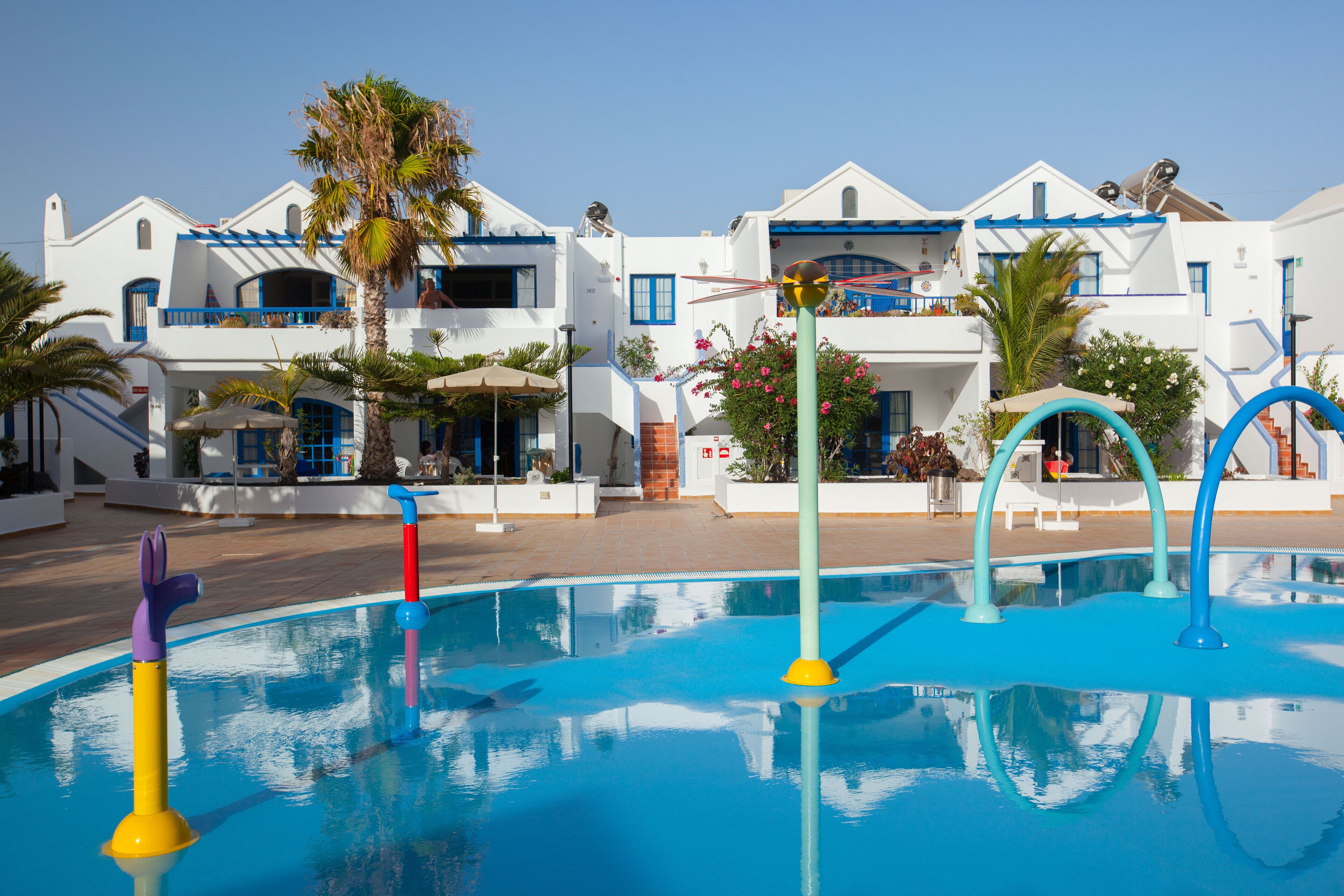 Area piscina con giochi colorati, una piscina blu cristallina e un edificio bianco con dettagli e balconi blu.