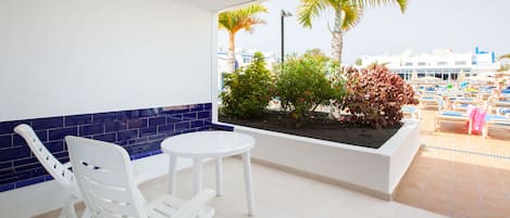 A balcony with white chairs and a table, a planter with plants, and a view of a beach with palm trees and a building in the background.