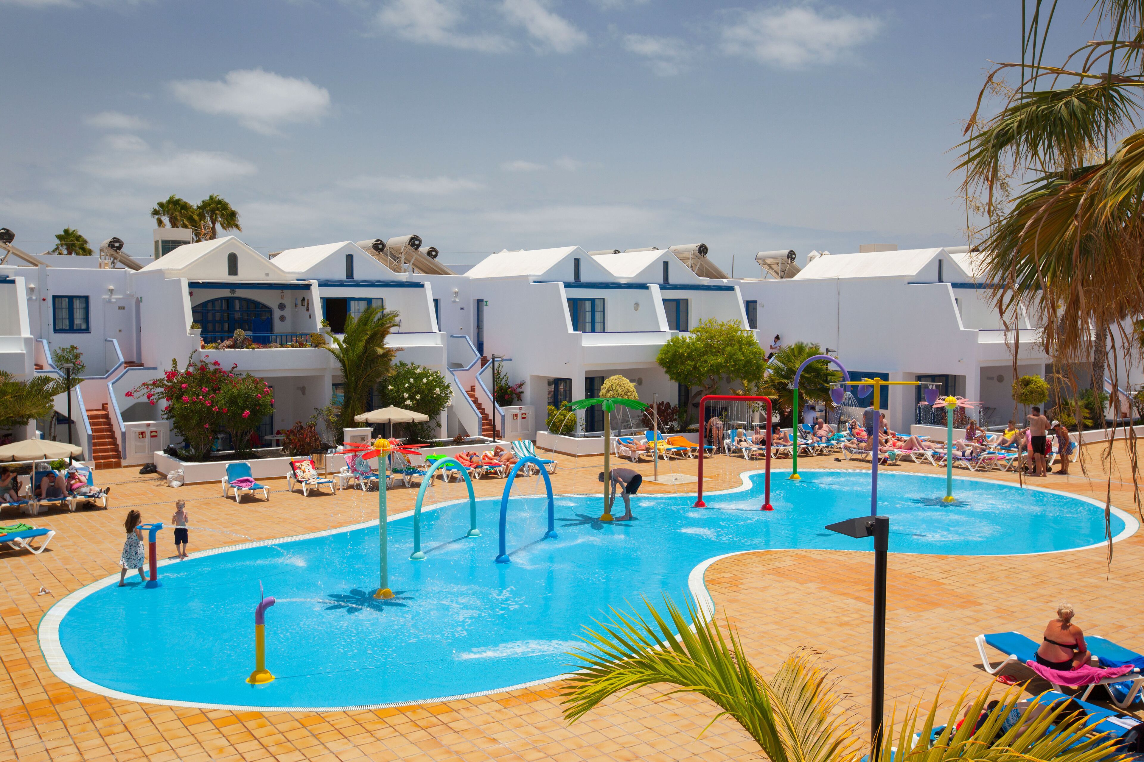 A swimming pool area with lounge chairs and umbrellas, surrounded by white buildings with blue accents.