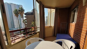 A balcony with a round table and chairs, a small bed, and a view of a cityscape with buildings and palm trees. - Europa Center (Benidorm)