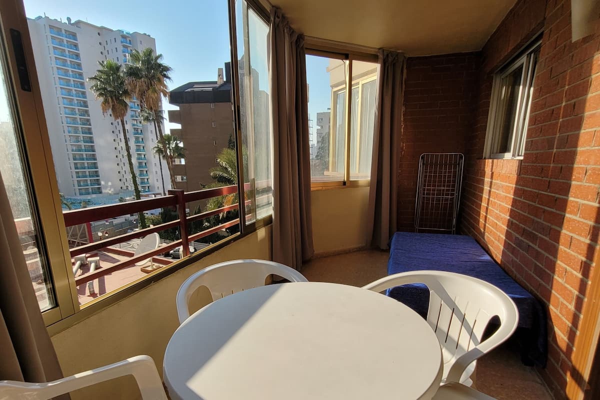 A balcony with a round table and chairs, a small bed, and a view of a cityscape with buildings and palm trees.