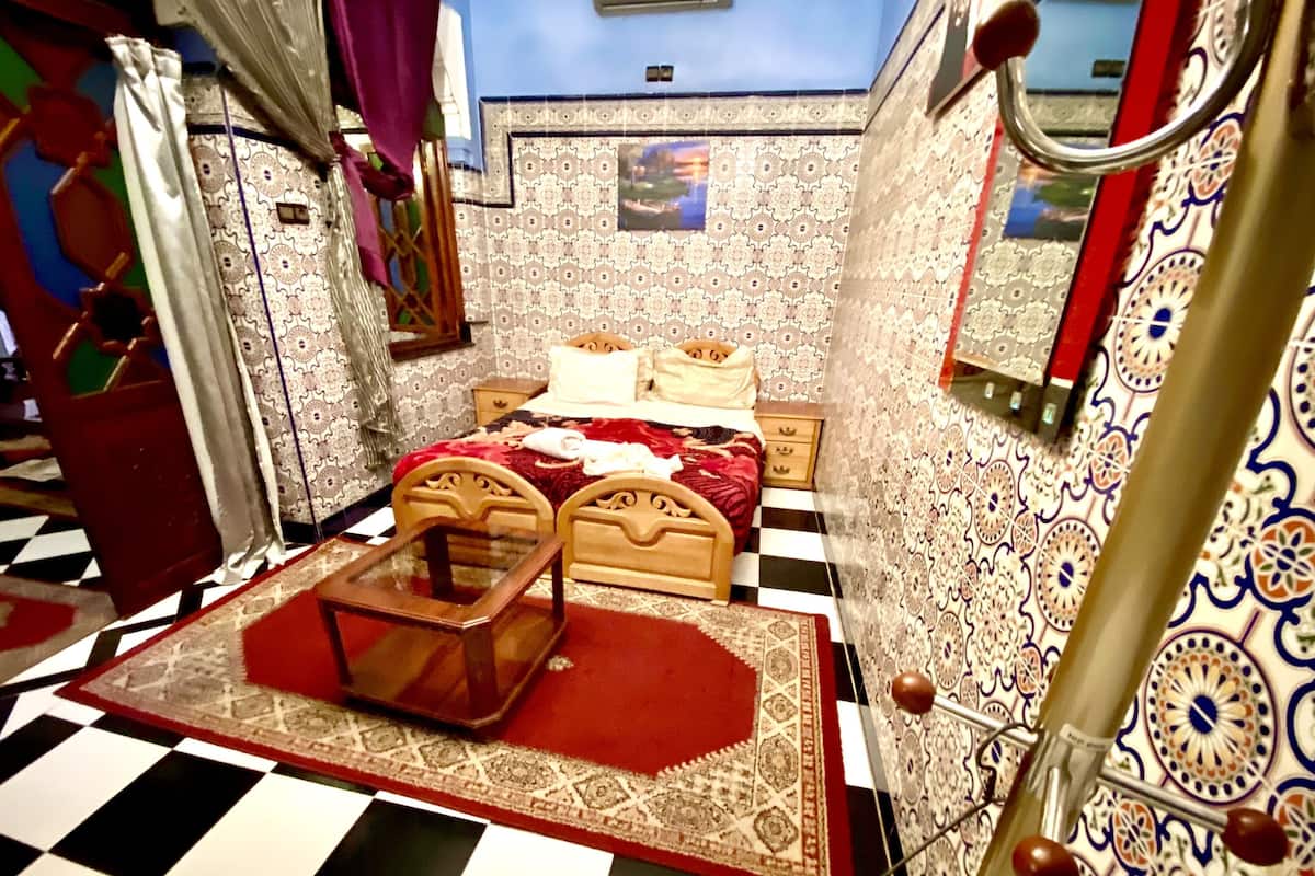 A room with a checkered floor, a bed with a red and white bedspread, a wooden headboard, a small table with a glass top, and patterned walls.