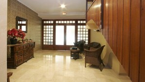 Un pasillo amplio con suelo de madera, una silla de mimbre, un mueble de madera con un jarrón de flores y una puerta de madera tradicional con paneles de vidrio.
