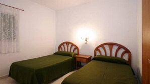 Una habitación pequeña y limpia con dos camas individuales, una mesita de noche, una lámpara de pared y una ventana con cortinas de gasa.