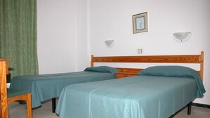 Una habitació amb dos llits individuals, una cadira de fusta, una taula petita i una cortina verda.