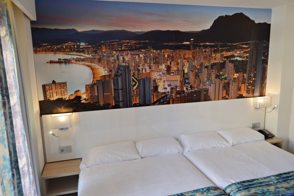 A hotel room with a large cityscape painting on the wall, a bed, and bedside lamps.